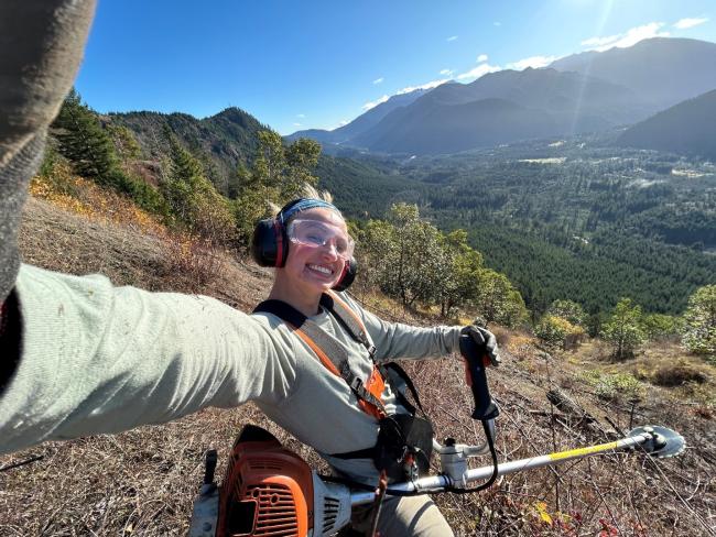 Aynslee McNeel, Washington Service Corps member serving at the Center for Natural Lands Management, uses a brush cutter to remove shrubs on land in the Olympia area. She's wearing goggles and headphones and is smiling while she takes a selfie.