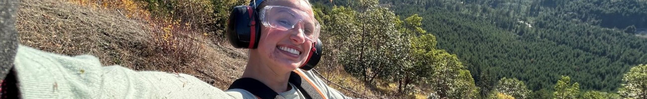 Aynslee McNeel, Washington Service Corps member serving at the Center for Natural Lands Management, uses a brush cutter to remove shrubs on land in the Olympia area. She's wearing goggles and headphones and is smiling while she takes a selfie.